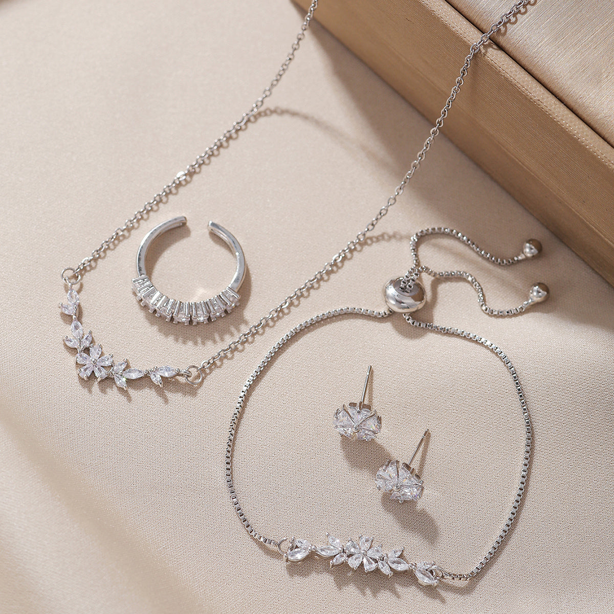 “Silver zircon jewelry set with necklace, bracelet, adjustable ring and earrings displayed on beige background”