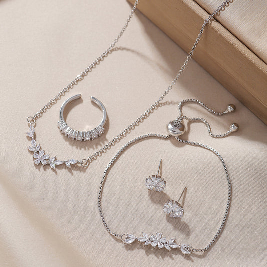 “Silver zircon jewelry set with necklace, bracelet, adjustable ring and earrings displayed on beige background”